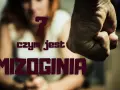 Zaciśnięta pięść i niewyraźna postać kobiety na tle. Pytanie "czym jest mizoginia" sugeruje przemoc i nienawiść wobec kobiet.