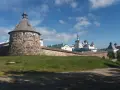 Kamienna wieża i mury obronne klasztoru na wyspach sołowieckich, z białymi cerkwiami i kopułami w tle, pod błękitnym niebem.