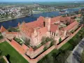Ogromny zamek krzyżacki w Polsce, z czerwonymi dachami i ceglanymi murami, otoczony zielenią i rzeką.