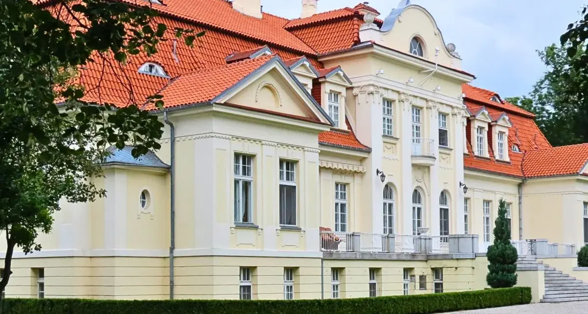 Dawny folwark szlachecki, dziś pałac z czerwoną dachówką, otoczony zielenią i brukowanym podjazdem.