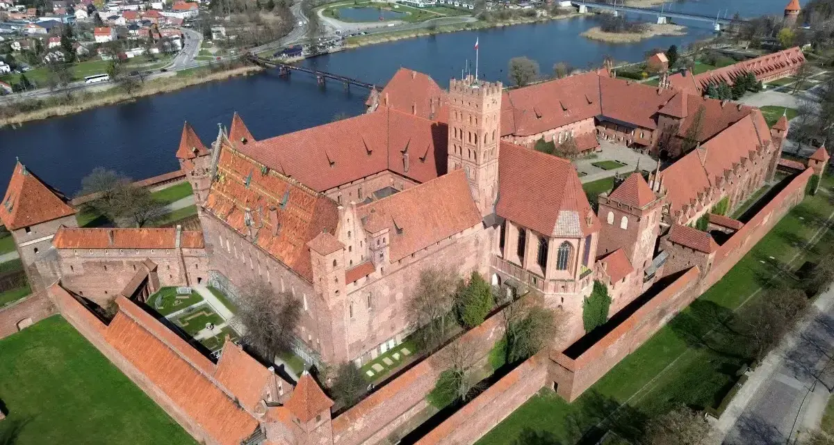 Ogromny zamek krzyżacki w Polsce, z czerwonymi dachami i ceglanymi murami, otoczony zielenią i rzeką.