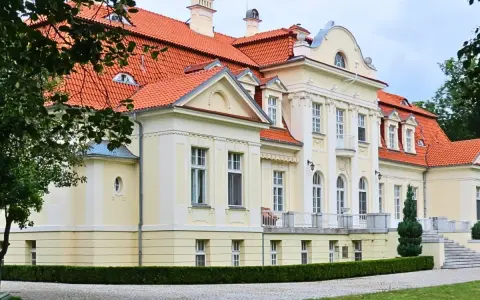 Dawny folwark szlachecki, dziś pałac z czerwoną dachówką, otoczony zielenią i brukowanym podjazdem.