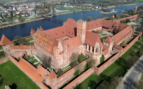 Ogromny zamek krzyżacki w Polsce, z czerwonymi dachami i ceglanymi murami, otoczony zielenią i rzeką.