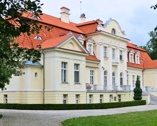 Dawny folwark szlachecki, dziś pałac z czerwoną dachówką, otoczony zielenią i brukowanym podjazdem.