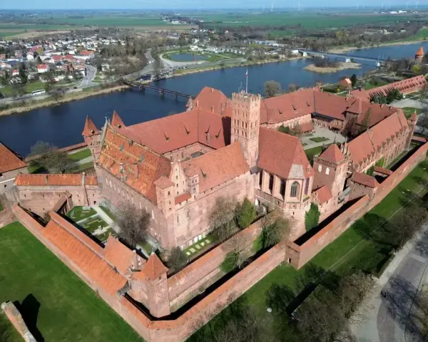 Ogromny zamek krzyżacki w Polsce, z czerwonymi dachami i ceglanymi murami, otoczony zielenią i rzeką.