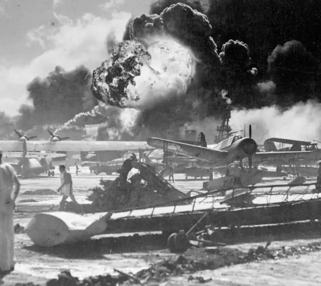 Dramatyczny wybuch w Pearl Harbor. Dym unosi się nad samolotami i budynkami, świadcząc o chaosie ataku.