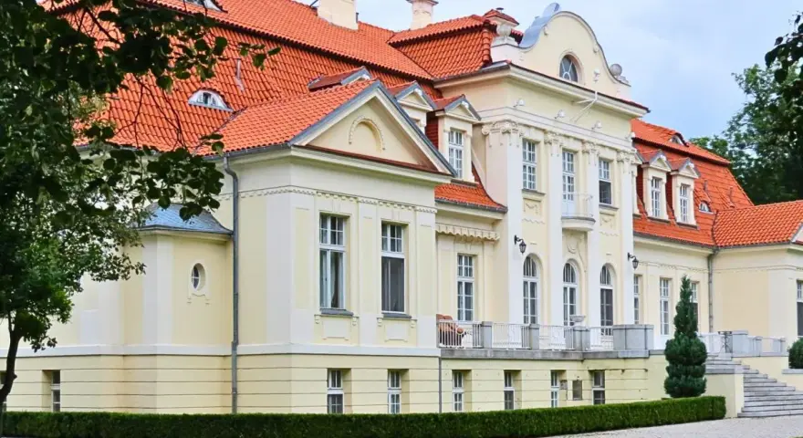 Dawny folwark szlachecki, dziś pałac z czerwoną dachówką, otoczony zielenią i brukowanym podjazdem.