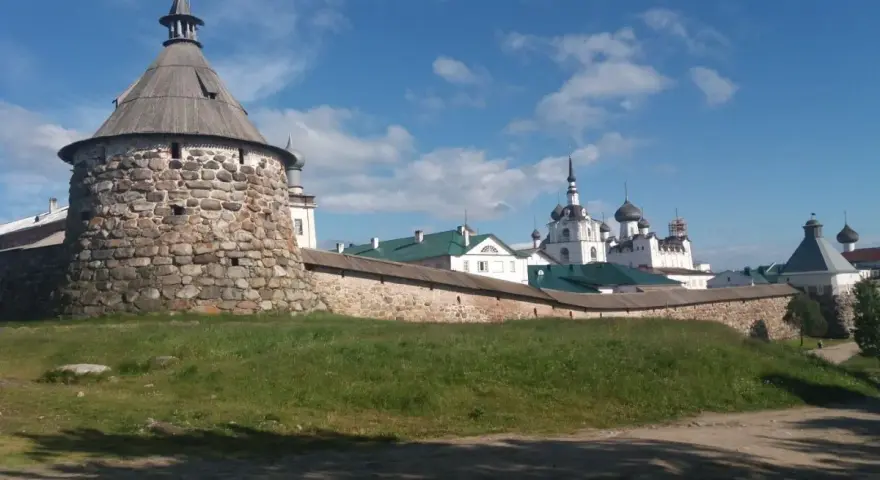 Kamienna wieża i mury obronne klasztoru na wyspach sołowieckich, z białymi cerkwiami i kopułami w tle, pod błękitnym niebem.