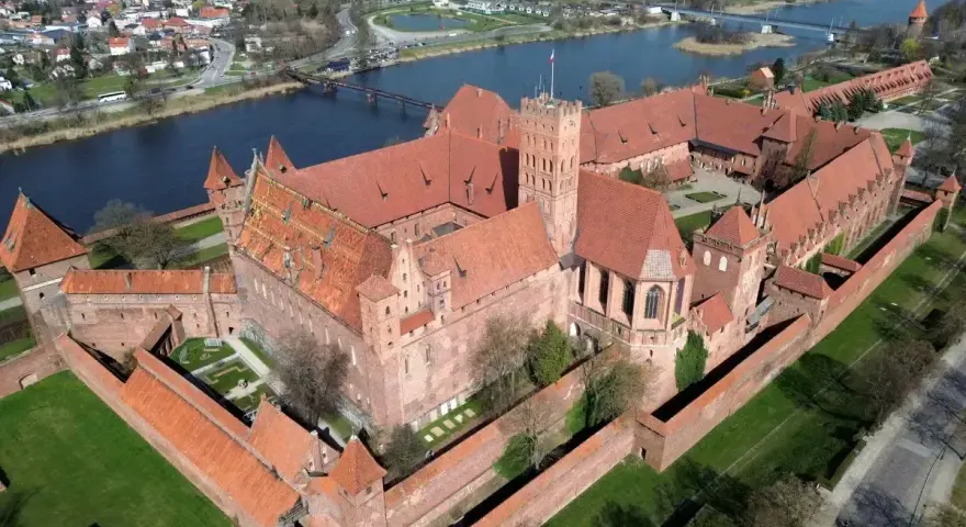 Ogromny zamek krzyżacki w Polsce, z czerwonymi dachami i ceglanymi murami, otoczony zielenią i rzeką.