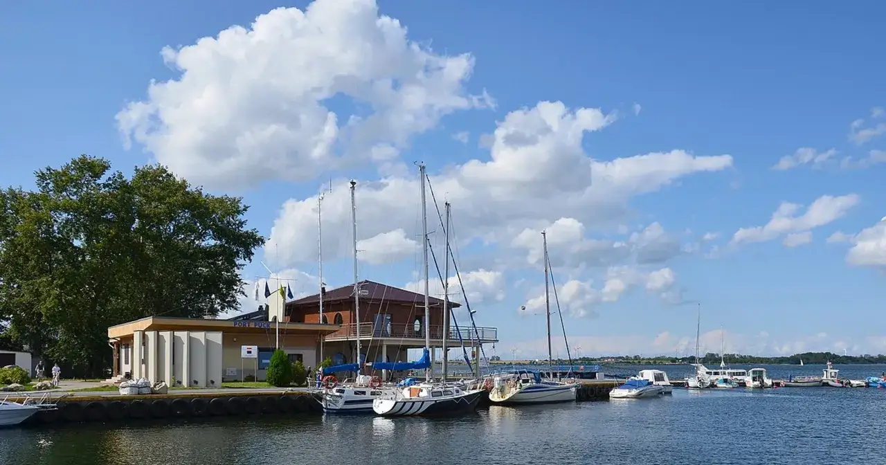 Port Puck z żagl&oacute;wkami przycumowanymi do nabrzeża. W tle budynek mariny i zielone drzewa.