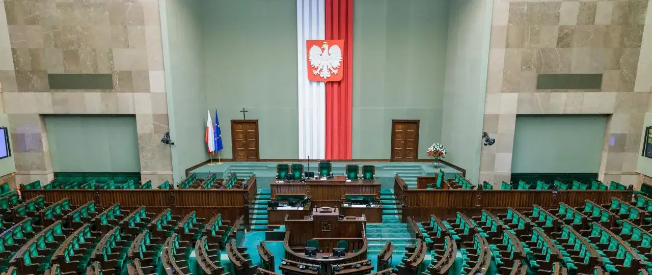 Sala sejmowa z rzędami zielonych foteli. Biało-czerwona flaga i godło Polski symbolizują co to suwerenność narodu.