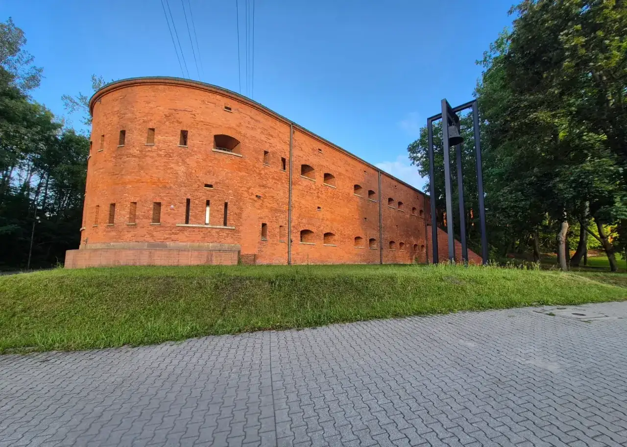 Ceglana twierdza Muzeum Katyńskiego w Warszawie, z charakterystycznymi otworami okiennymi, otoczona zielenią.