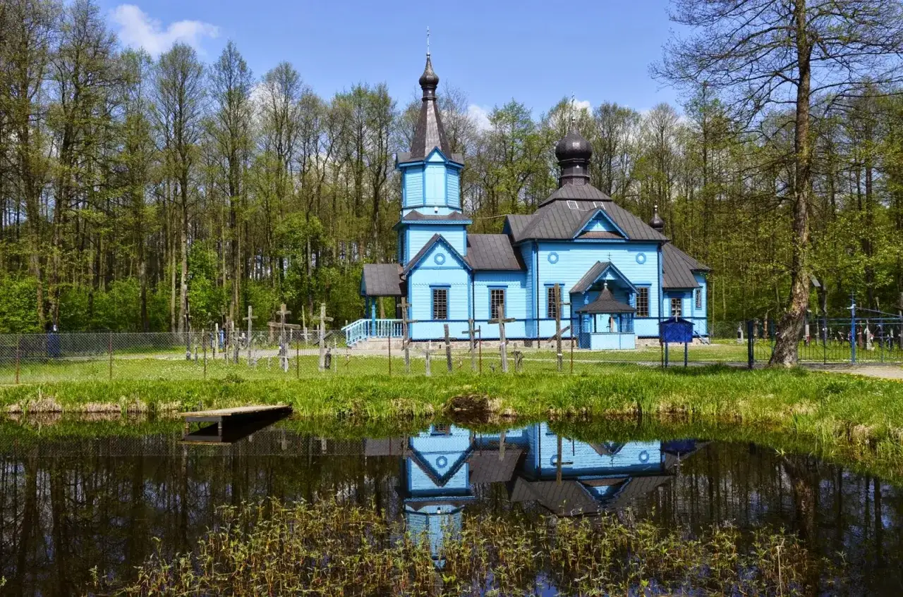 Niebieska cerkiew na Podlasiu, odbijająca się w spokojnej wodzie, otoczona lasem i cmentarzem z krzyżami.