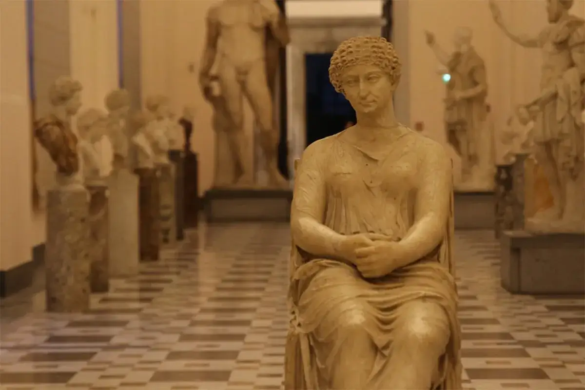 Kamienna postać siedzi w muzeum archeologicznym Neapol, otoczona innymi rzeźbami.